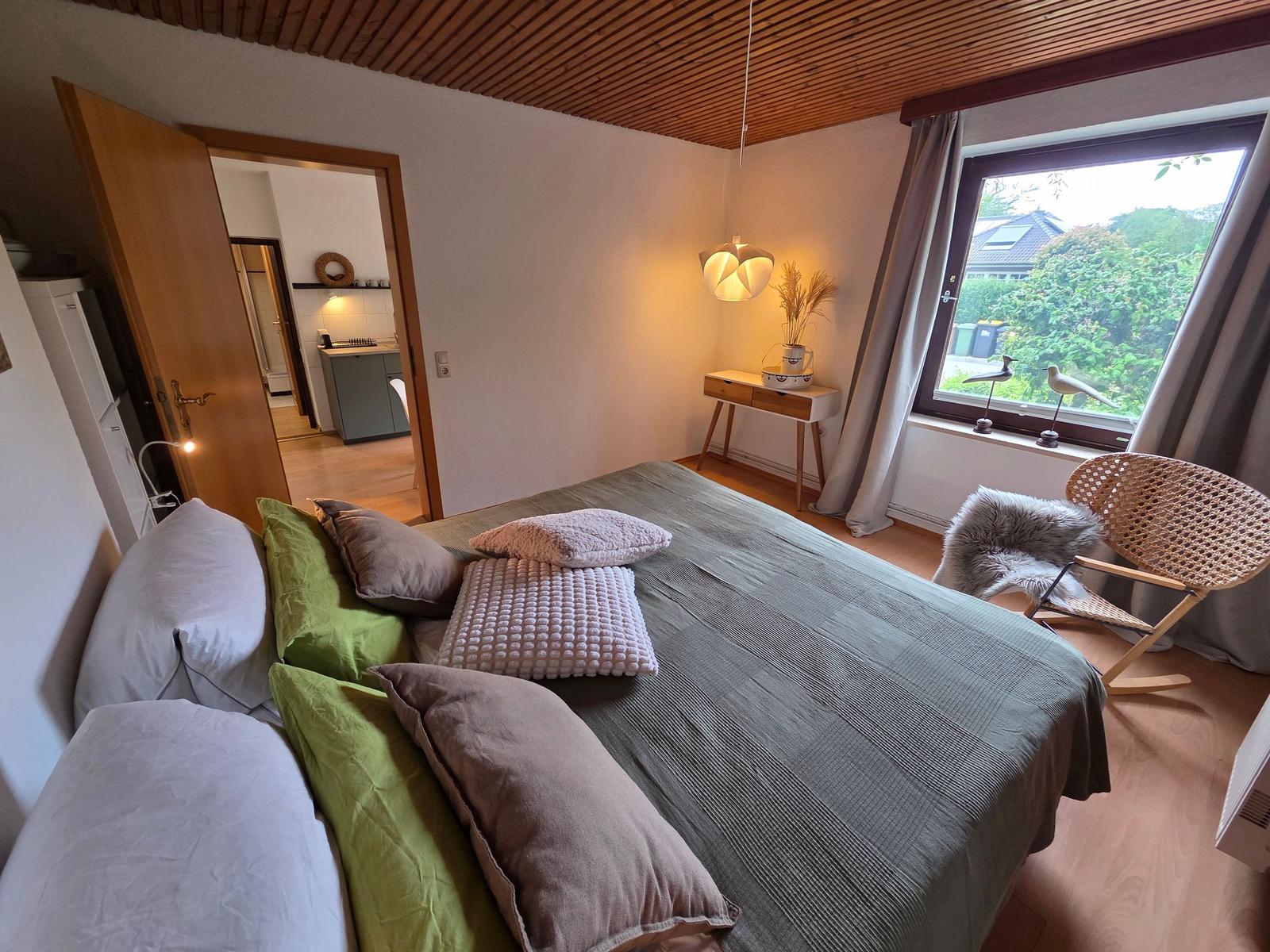 Schlafzimmer mit Bett, Stuhl und Fenster. Durchgang zur Küche.