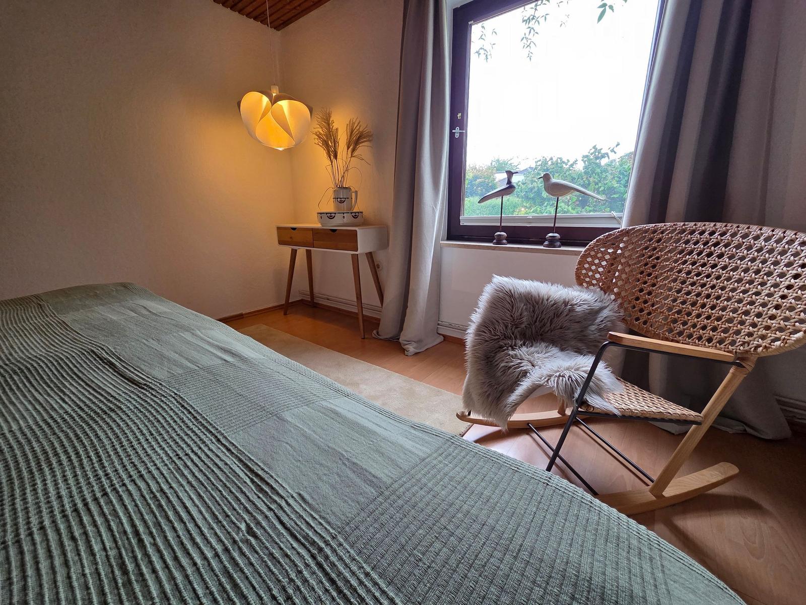 Schlafzimmer mit Bett, Schaukelstuhl und Fenster mit Blick auf Grün.