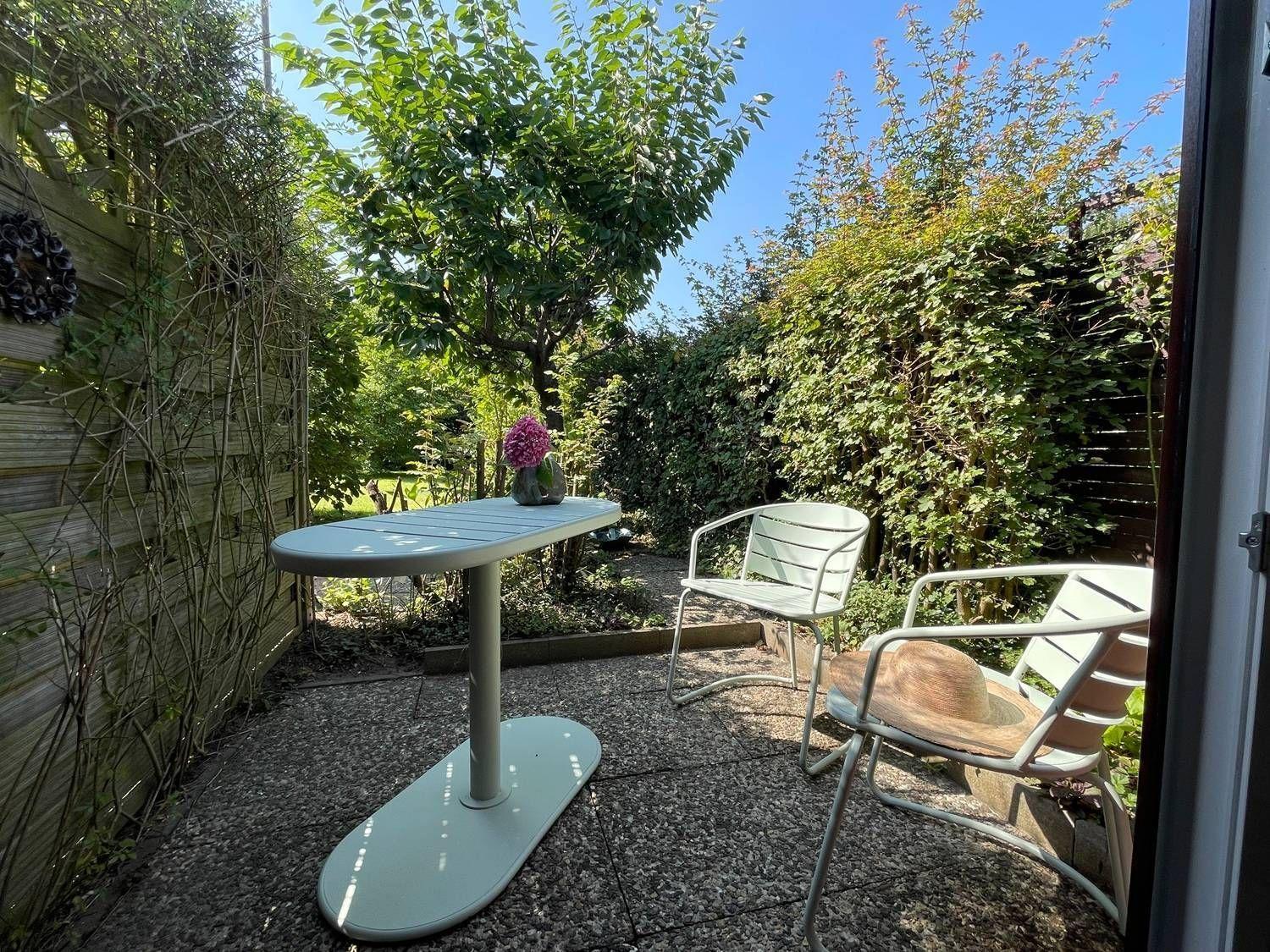 Terrasse mit Tisch und zwei Stühlen, umgeben von Grün und einem Holzzaun.