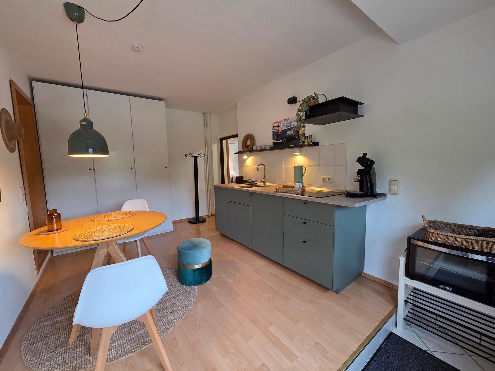 Küche mit Essbereich, Holzboden und grauen Schränken. Tisch mit Stühlen und Deckenlampe.