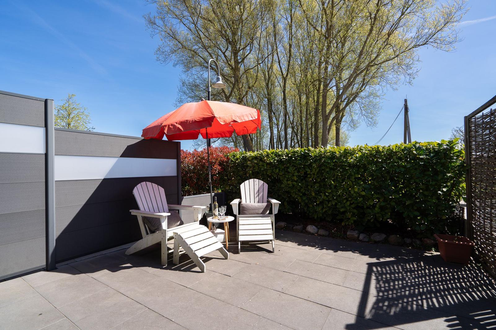 Terrasse mit zwei weißen Sitzgelegenheiten und rotem Sonnenschirm. Hintergrund: grüner Heckenzaun und Bäume.