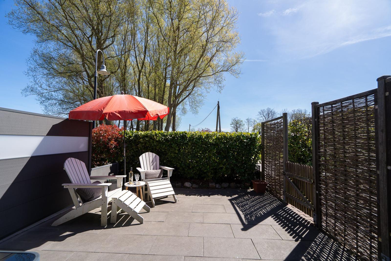 Terrasse mit zwei weißen Liegestühlen, rotem Sonnenschirm und Tisch unter Bäumen.