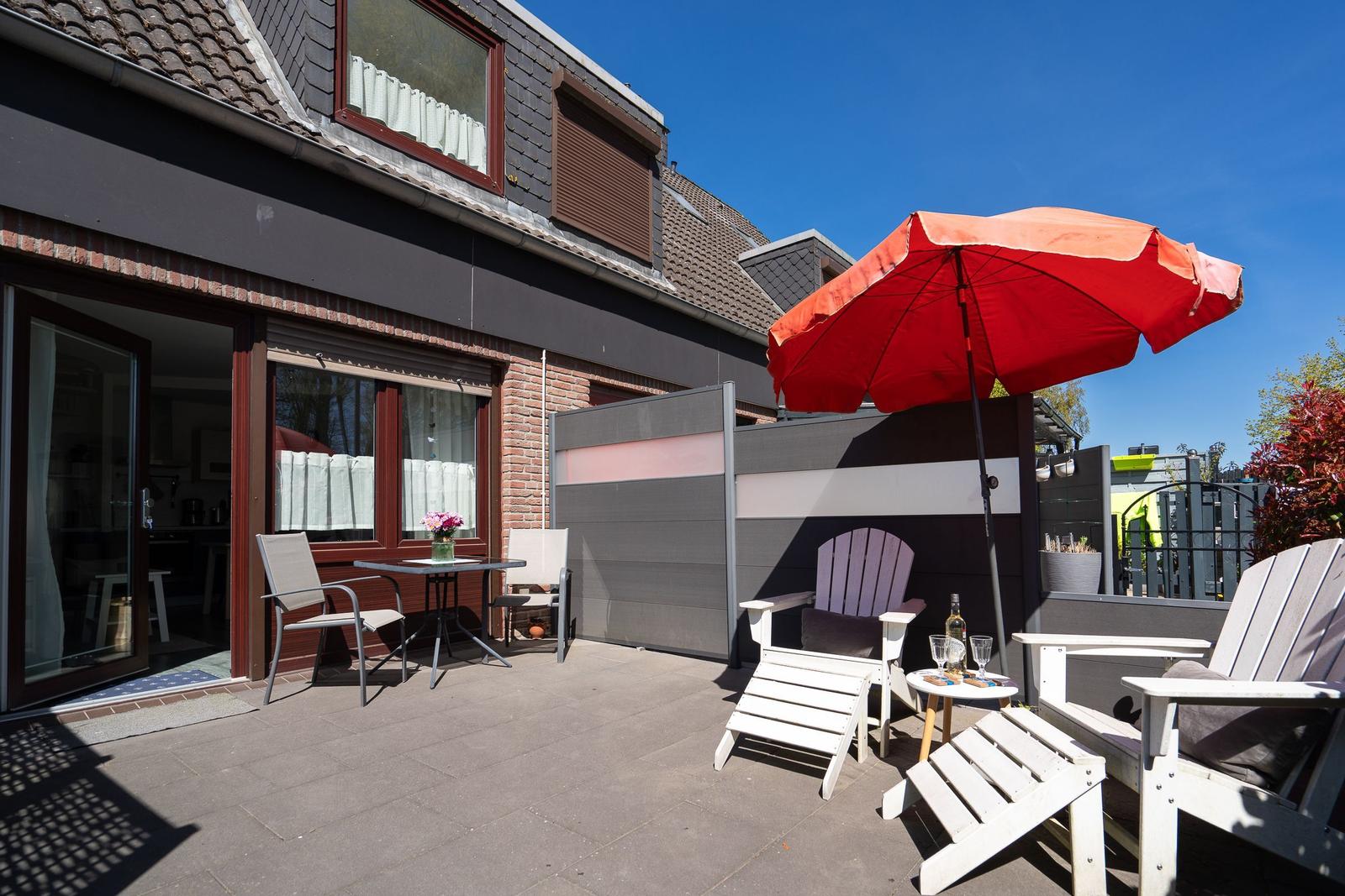 Terrasse mit Tisch, Stühlen und rotem Sonnenschirm. Haus mit Fenstern und Terrassentür.