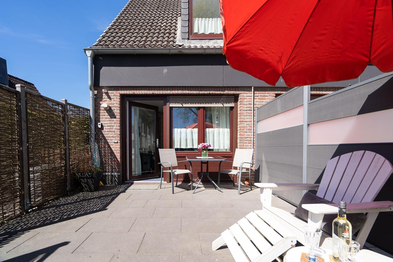 Terrasse mit Tisch, Stühlen und rotem Sonnenschirm. Hinterhof mit Zaun und Hausfassade.