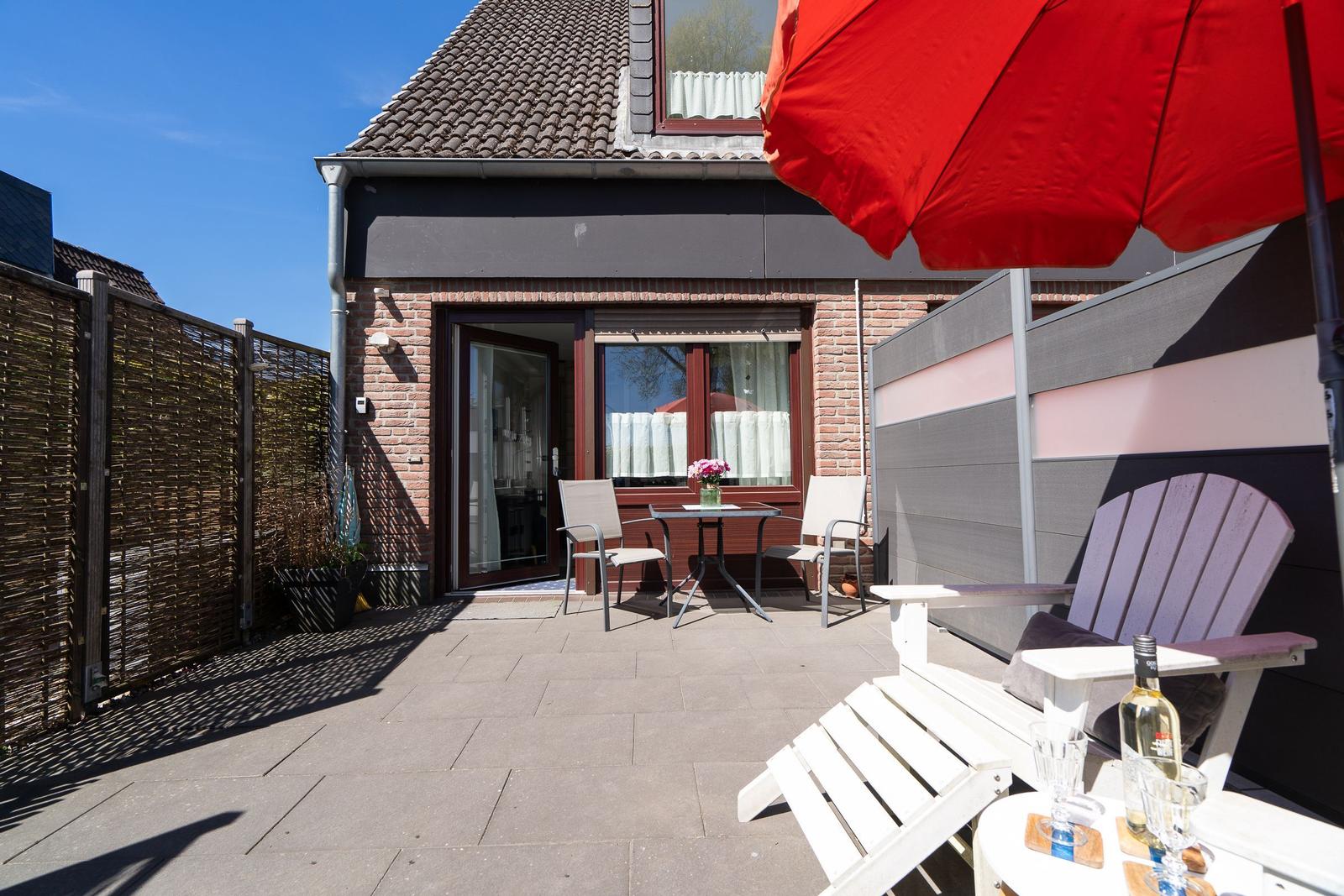 Terrasse mit Tisch, Stühlen und rotem Sonnenschirm. Hinterhof mit Zaun und Eingangstür.