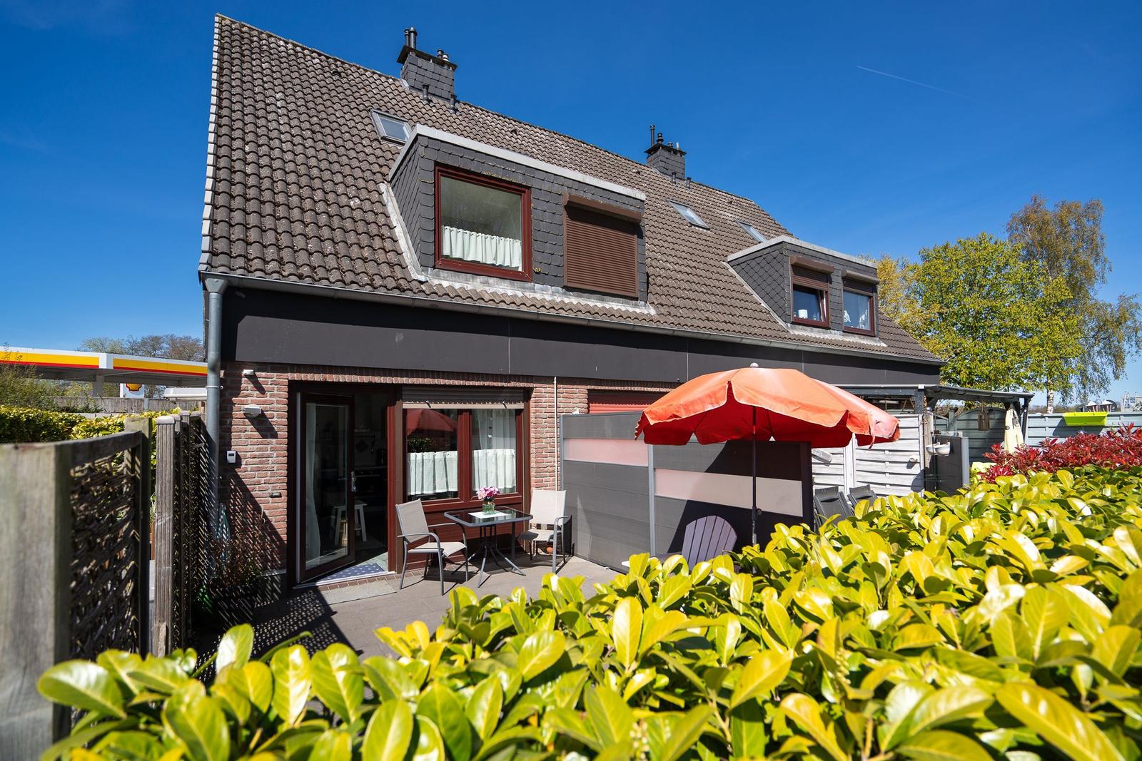 Haus mit Terrasse, rotem Sonnenschirm und grünen Sträuchern im Vordergrund.
