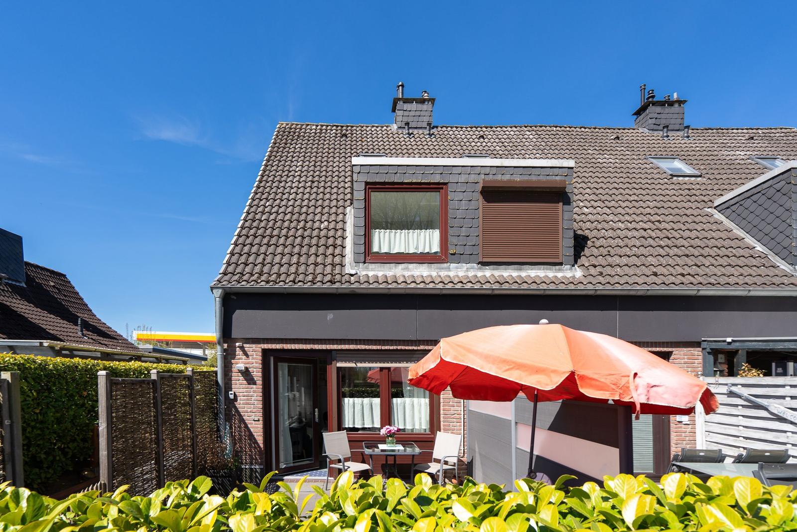 Haus mit Terrasse, orangefarbener Sonnenschirm und grüner Hecke unter blauem Himmel.