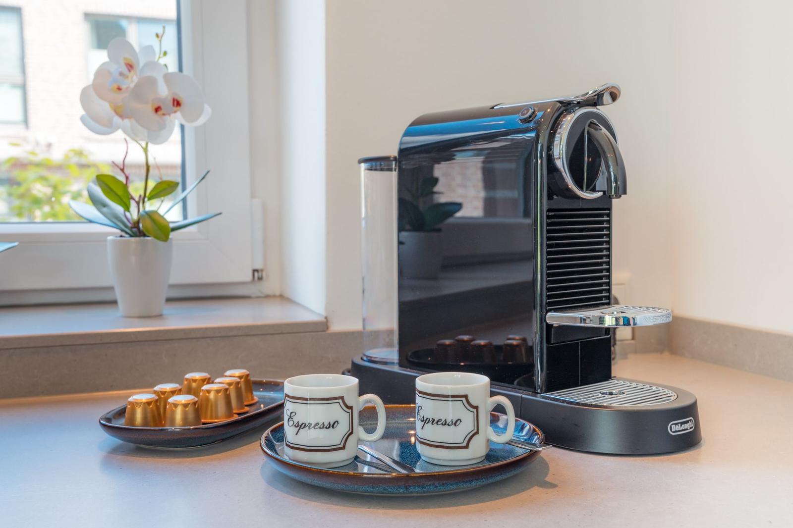 Espressomaschine mit Tassen und Kapseln auf Küchenarbeitsfläche neben Fenster mit Orchidee.