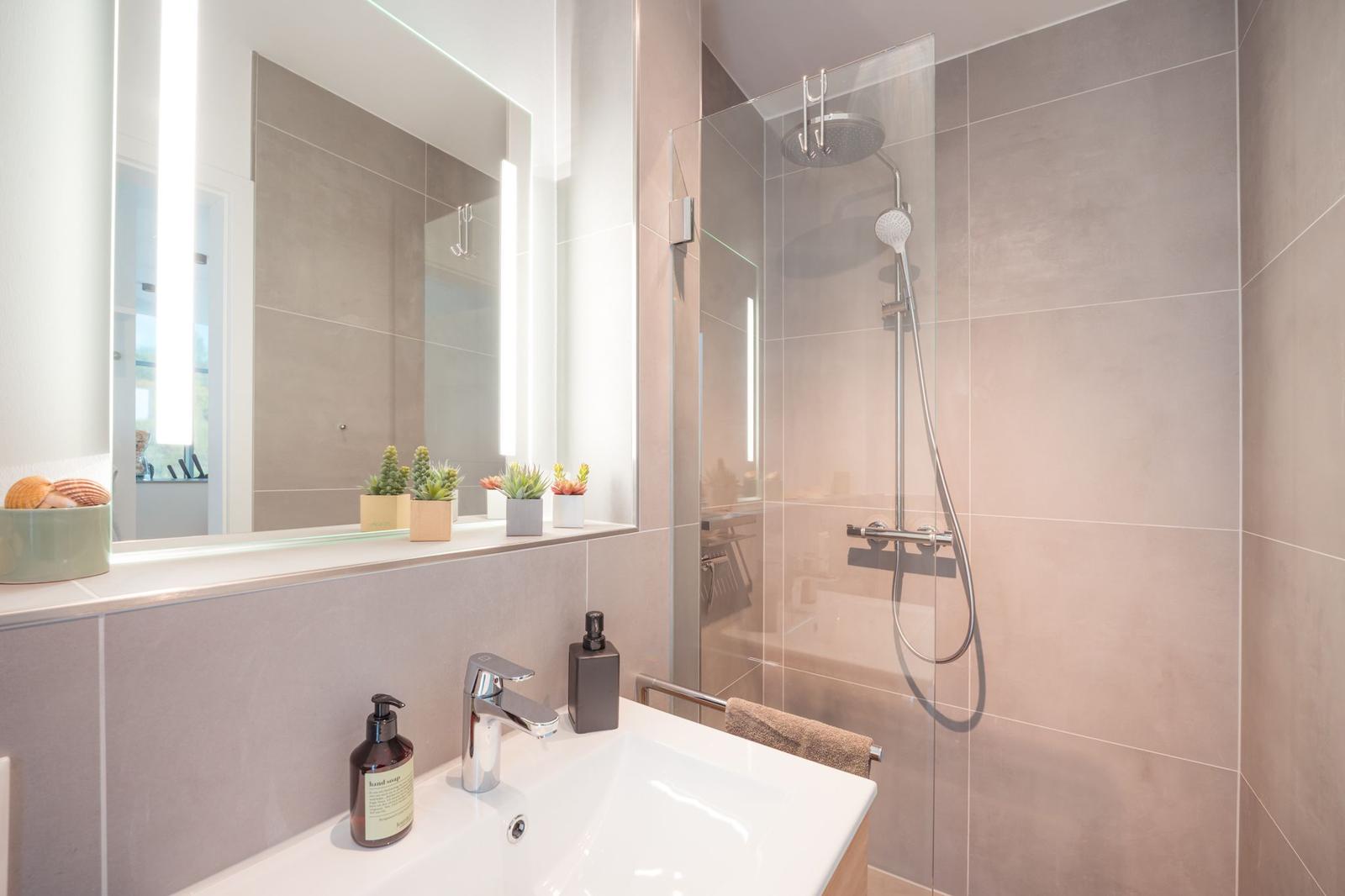 Badezimmer mit Dusche, Waschbecken und beleuchtetem Spiegel