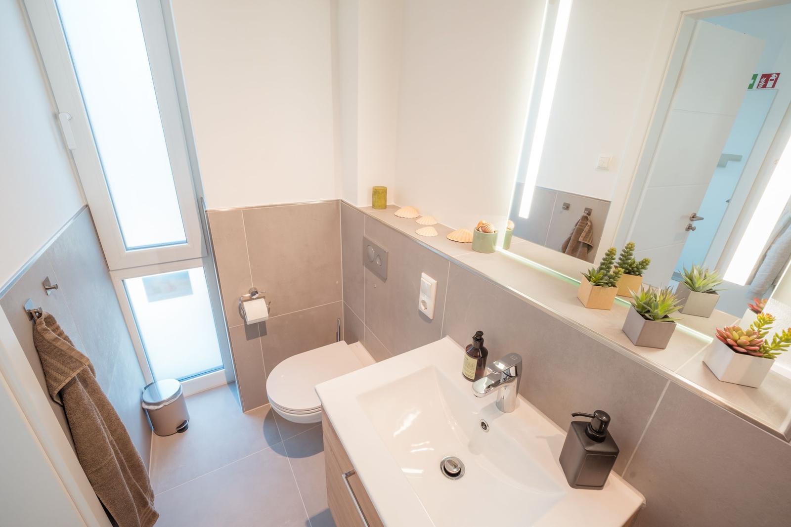 Badezimmer mit Waschbecken, Spiegel, Toilette und Fenster.