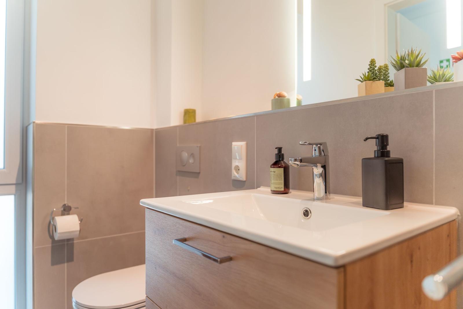 Bad mit Waschbecken, Spiegel und Toilette. Auf dem Waschtisch stehen Seifen- und Flüssigseifendosen.