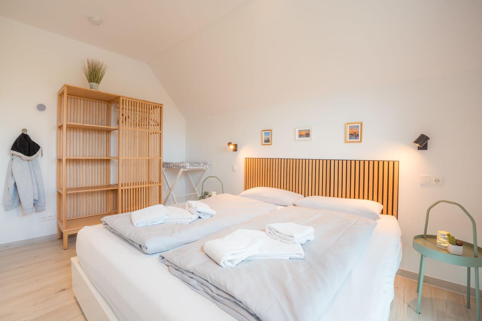Schlafzimmer mit Doppelbett, Holzregal und Nachttisch. An der Wand hängen Bilder und Lampen.