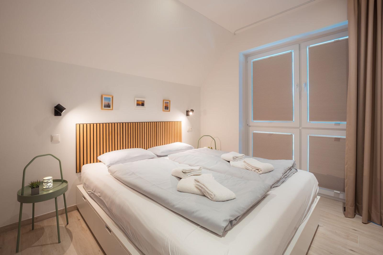 Schlafzimmer mit Doppelbett, Holzkopfteil und Fenster mit Jalousien.