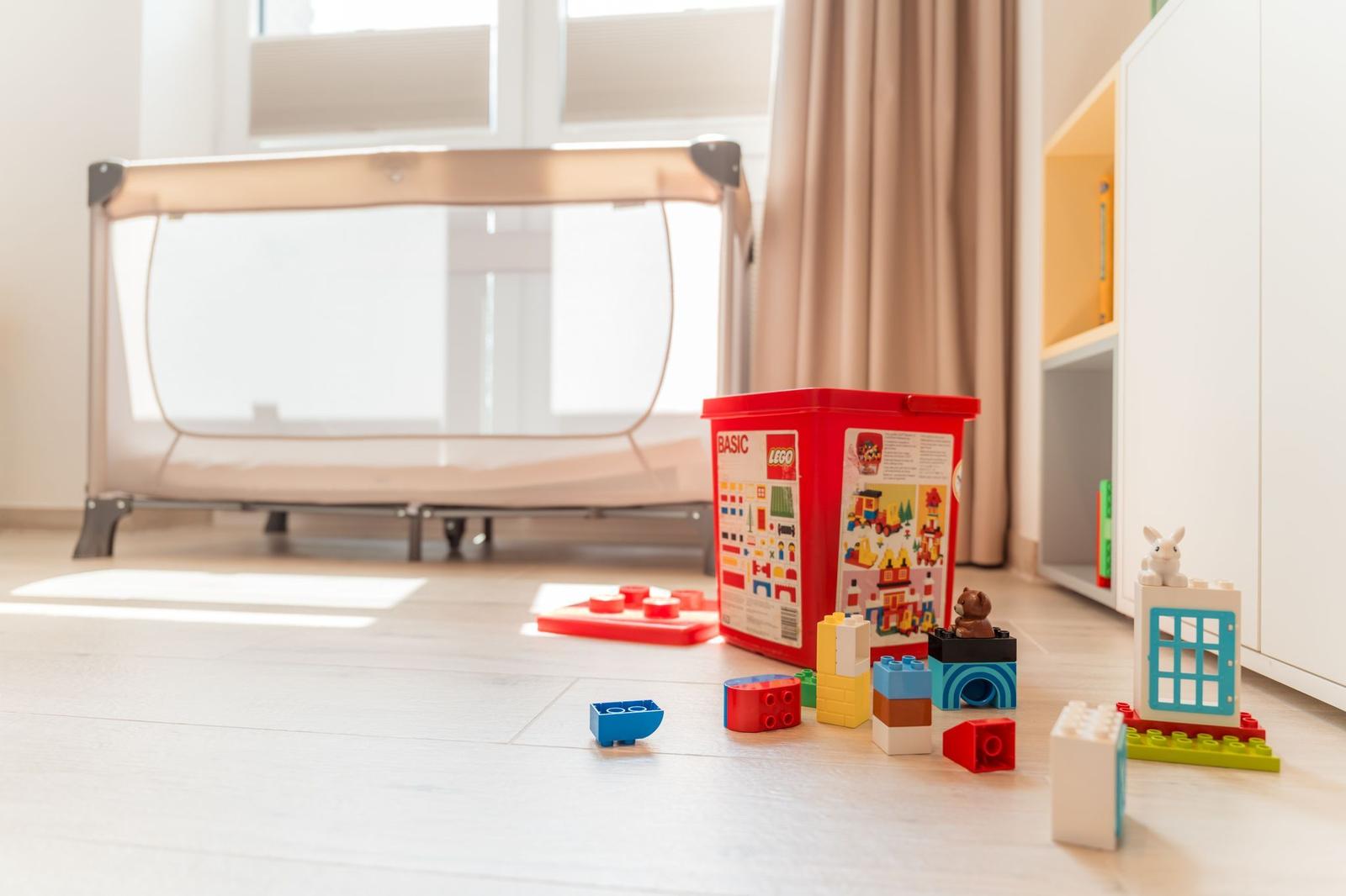 Kinderzimmer mit Spielzeugbox und Babybett. Holzboden und Fenster mit Jalousie.