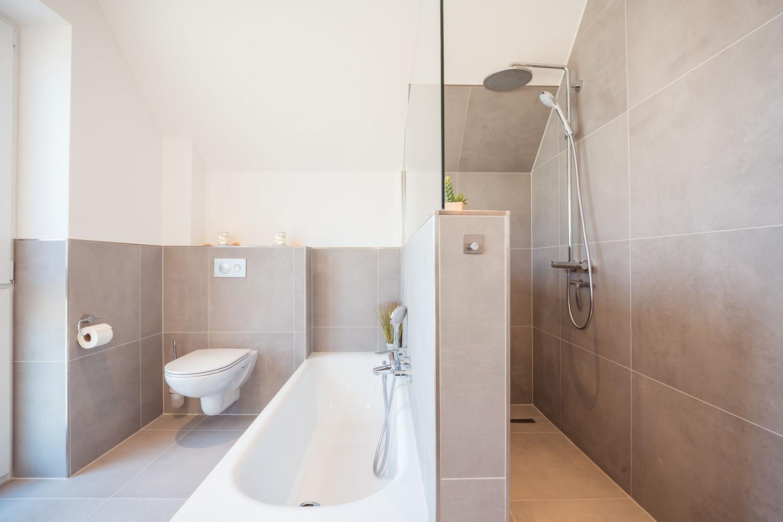 Bad mit Badewanne, Dusche und WC. Wandverkleidung aus grauen Fliesen.