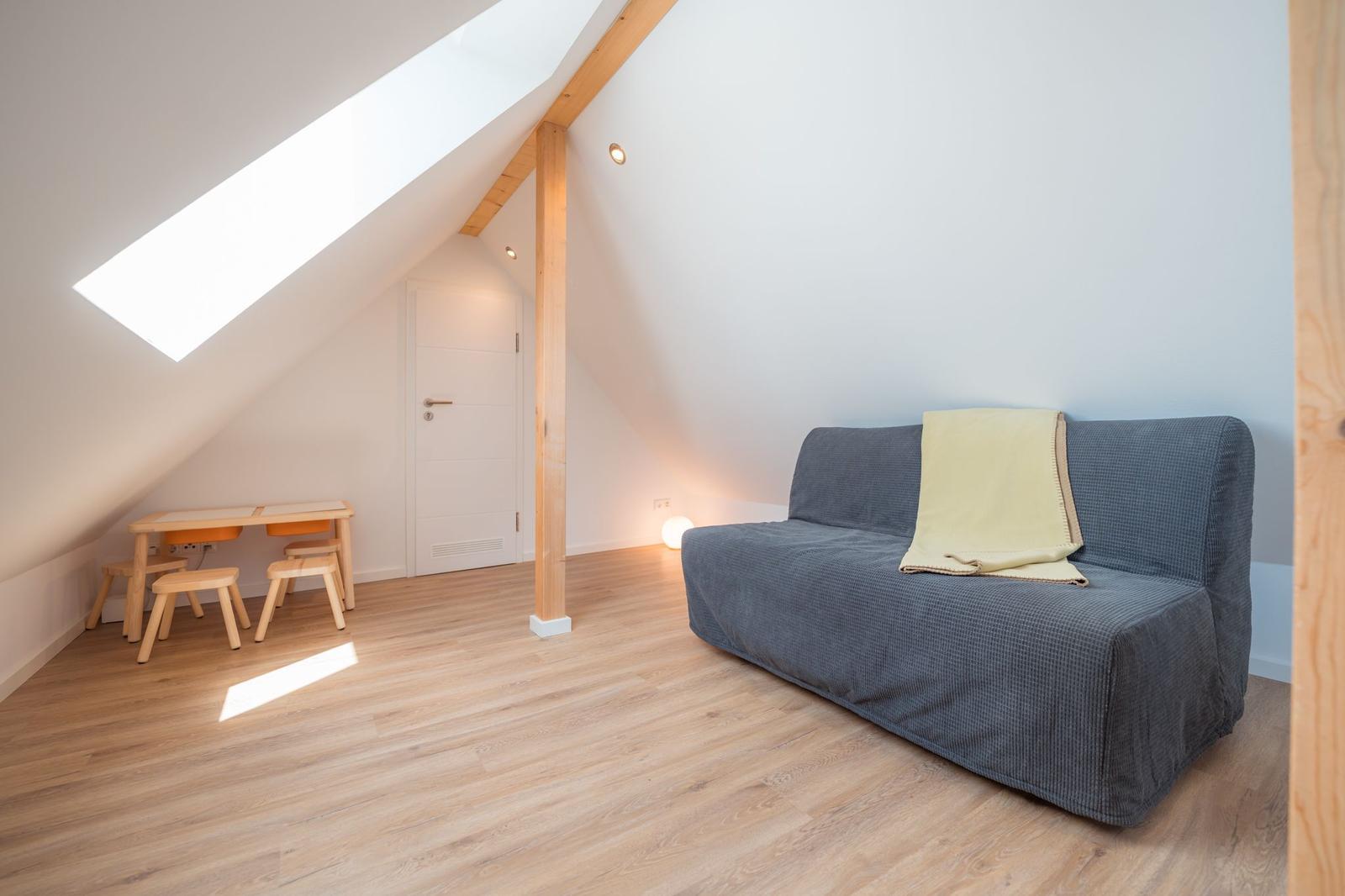 Dachgeschosszimmer mit Sofa, Schreibtisch und Holzboden. Dachfenster und Holzstütze sichtbar.