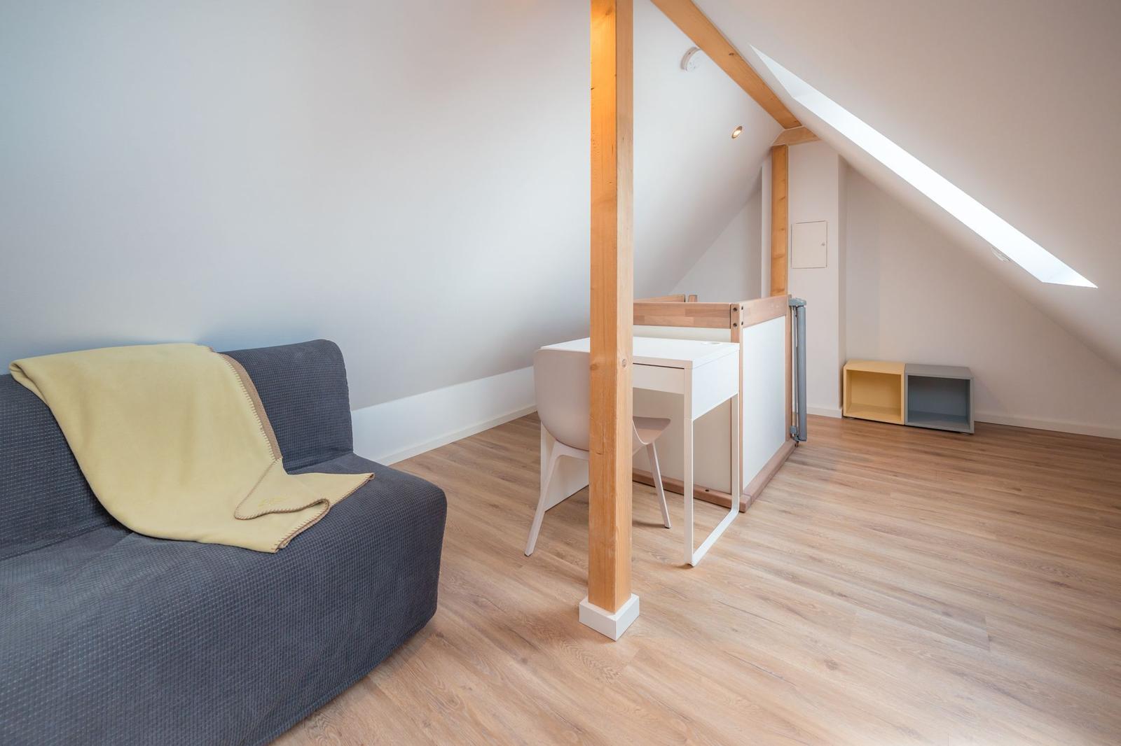 Ein Raum mit Sofa, Schreibtisch und Dachfenster. Holzdielenboden und Holzstützen.