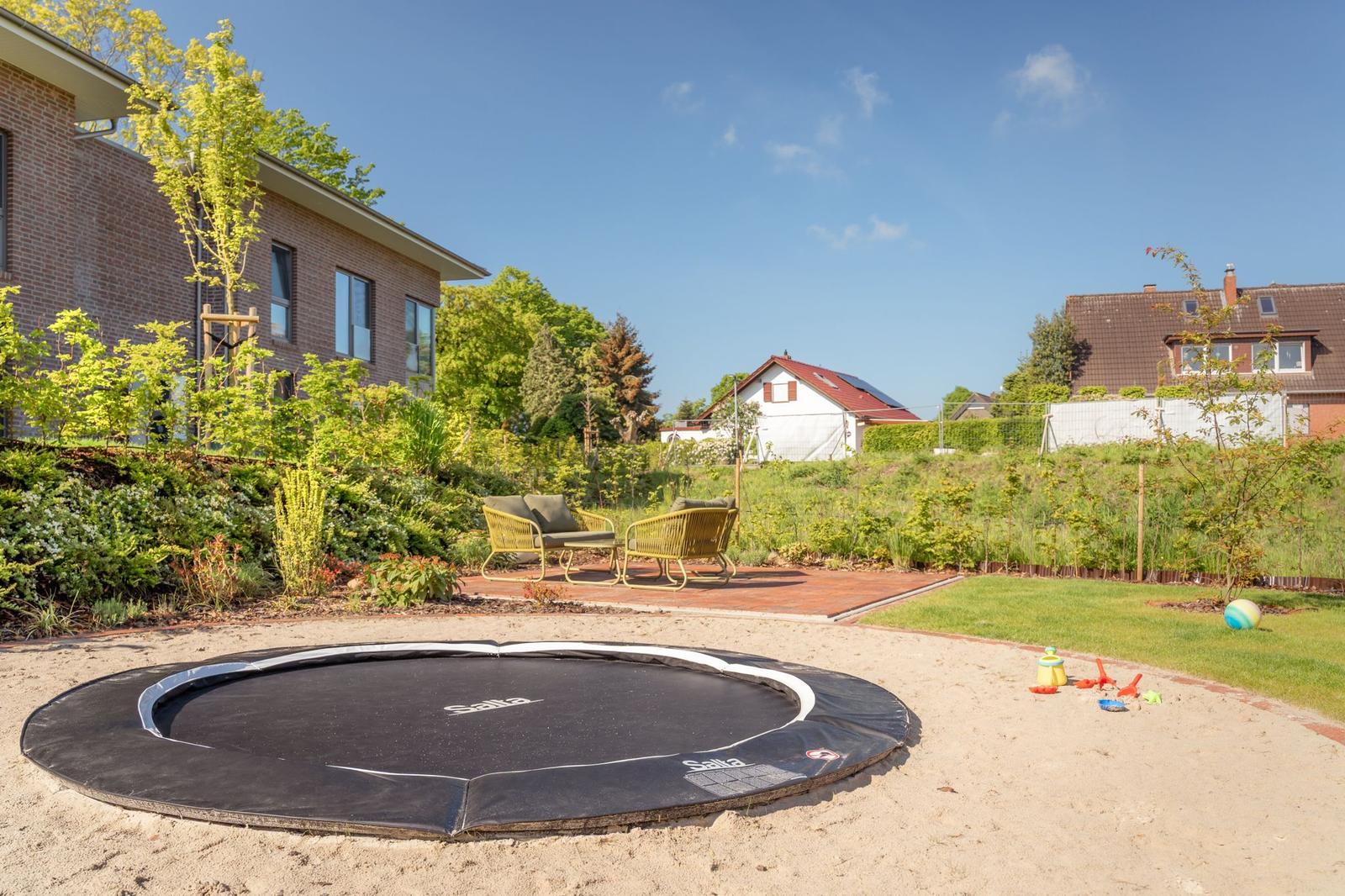 Trampolin im Sand, Gartenbereich mit Sitzgelegenheit und Blick auf Wohnhäuser.