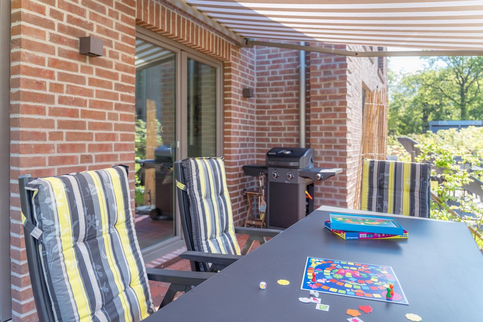Terrasse mit Tisch, Stühlen und Grill unter Markise. Spiel auf Tisch.