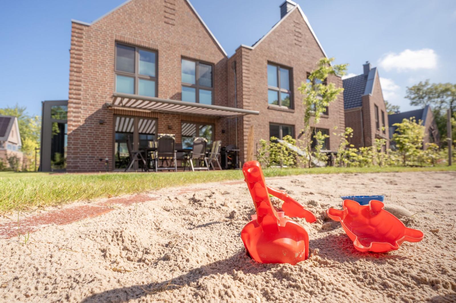Roter Sandkasten und Schaufel liegen im Sand vor einem Backsteinhaus mit Terrasse.