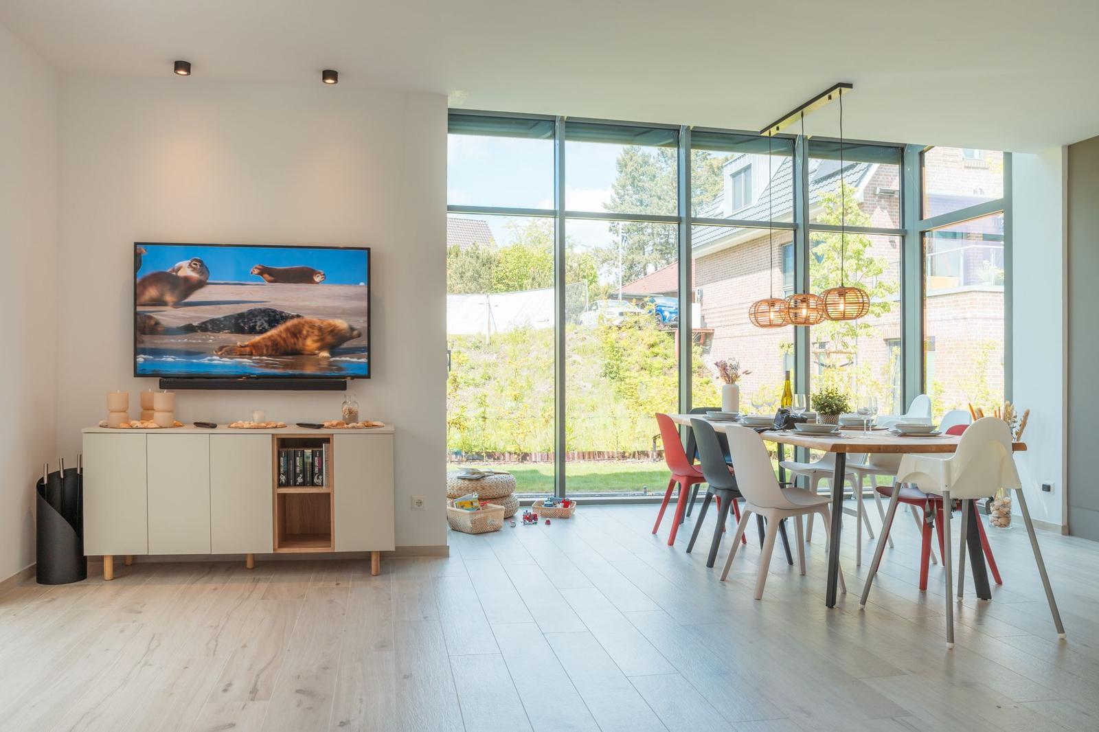 Wohn- und Essbereich mit TV, Esszimmertisch und großen Fenstern.