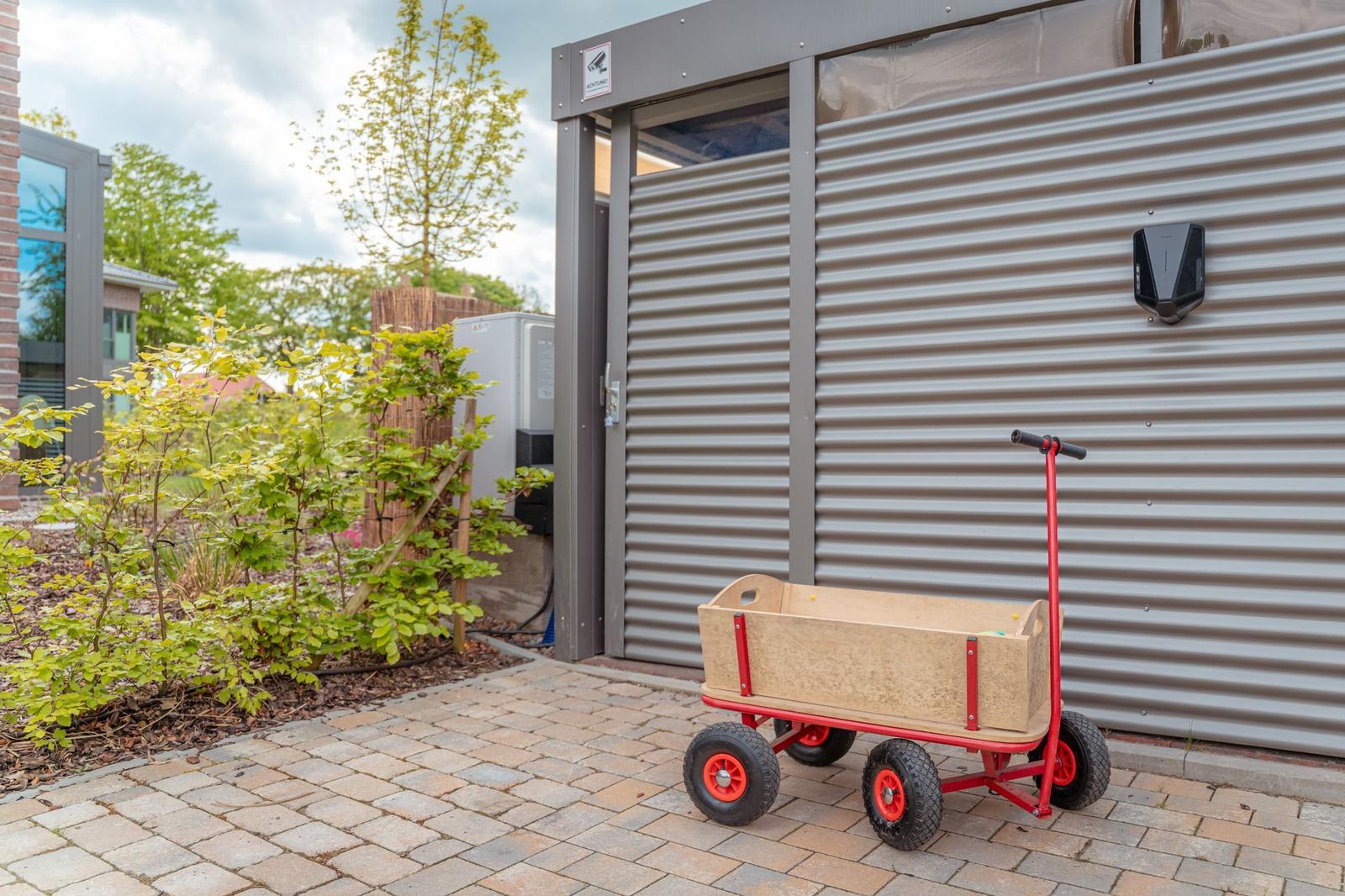Ein roter Kinderwagen steht vor einer Metallgarage auf einem gepflasterten Hof.