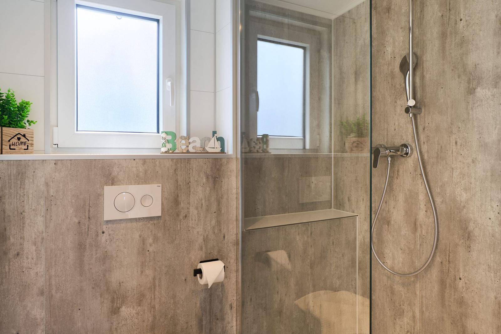 Dusche mit Glaswand und Toilette. Fenster mit Blick. Dekoration auf Fensterbank.