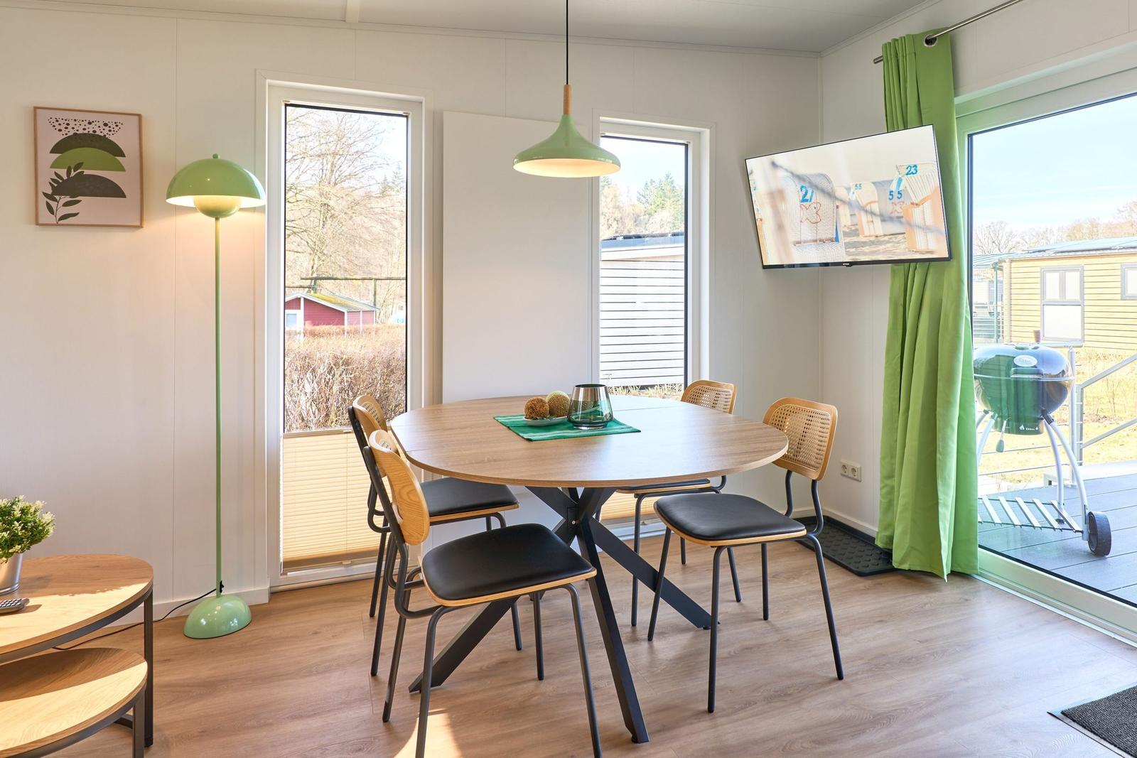 Esszimmer mit runden Tisch, vier Stühlen und großen Fenstern. Grüne Lampe und TV an der Wand.