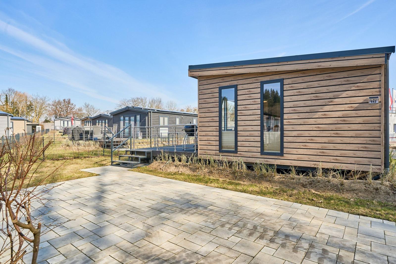 Holzhaus mit Terrasse und Grillplatz. Hintergrund: weitere Unterkünfte und Bäume.