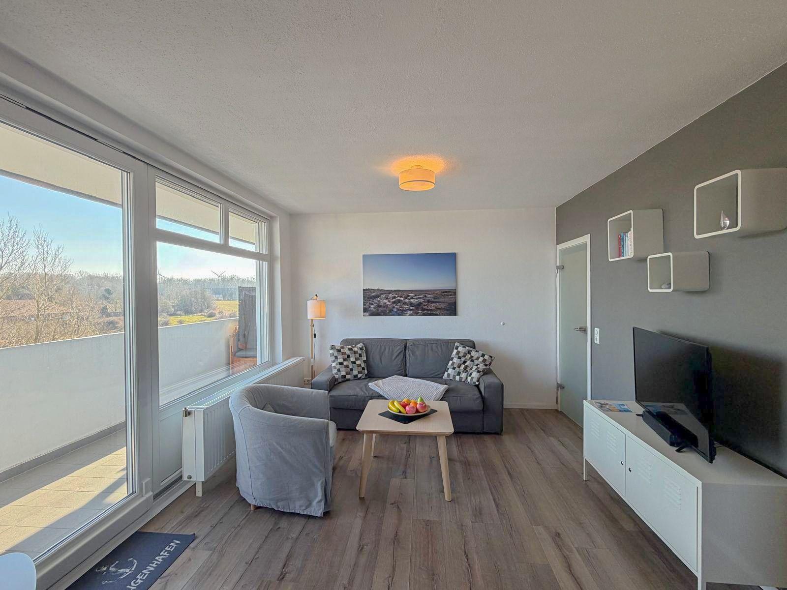 Wohnzimmer mit Sofa, Sessel, Couchtisch und TV. Große Fenster mit Blick auf Balkon.