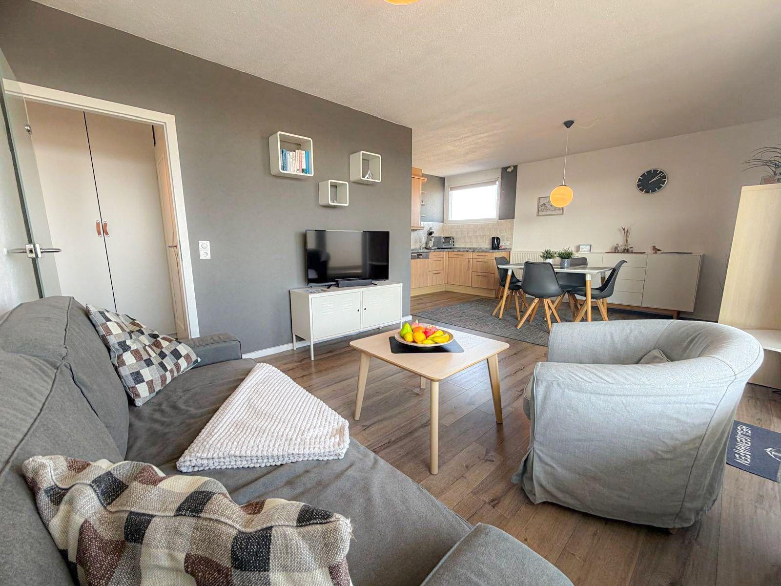 Wohnzimmer mit Sofa, Sessel und TV. Küche und Essbereich im Hintergrund.