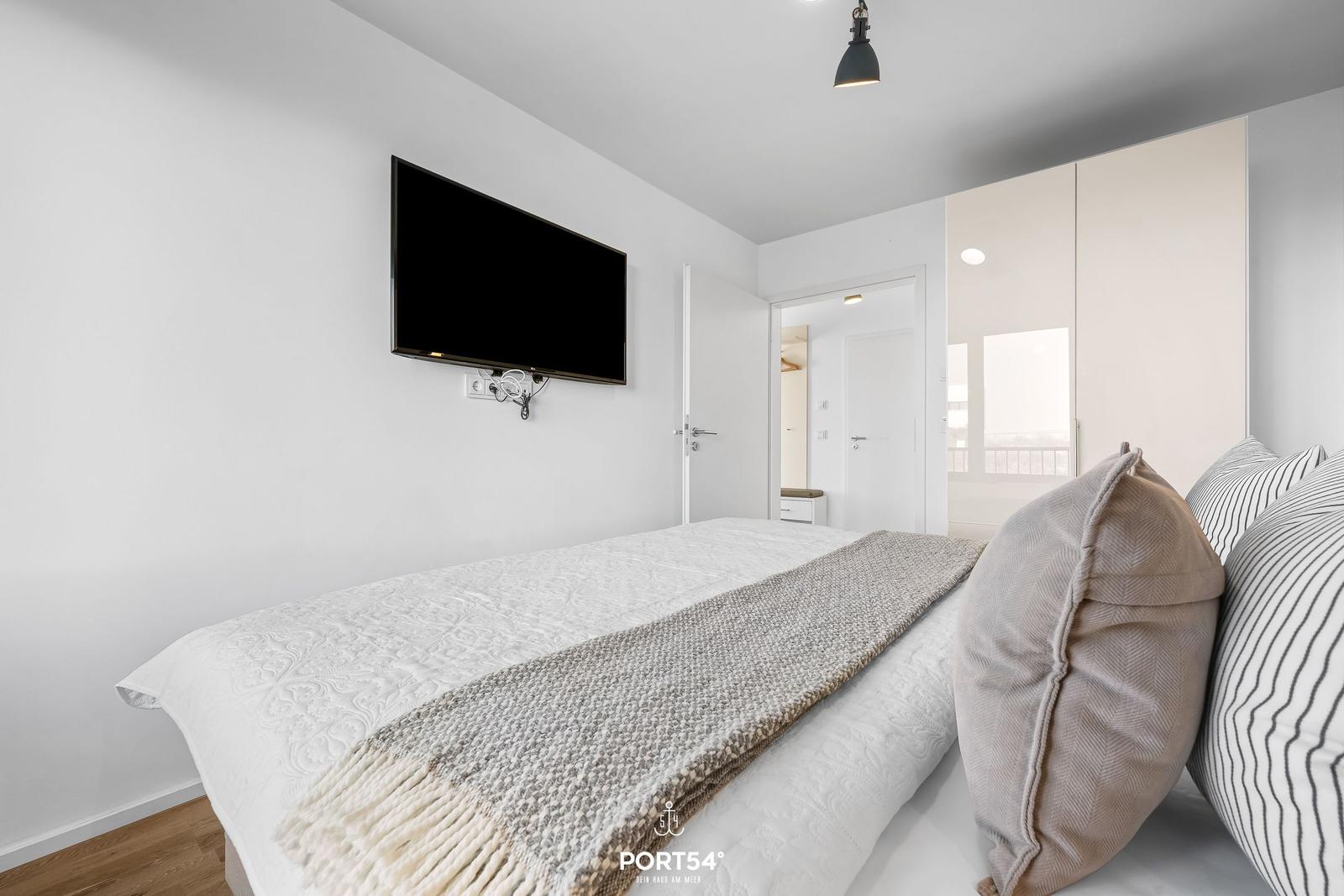 Schlafzimmer mit Bett, Fernseher an der Wand und Blick in den Flur.