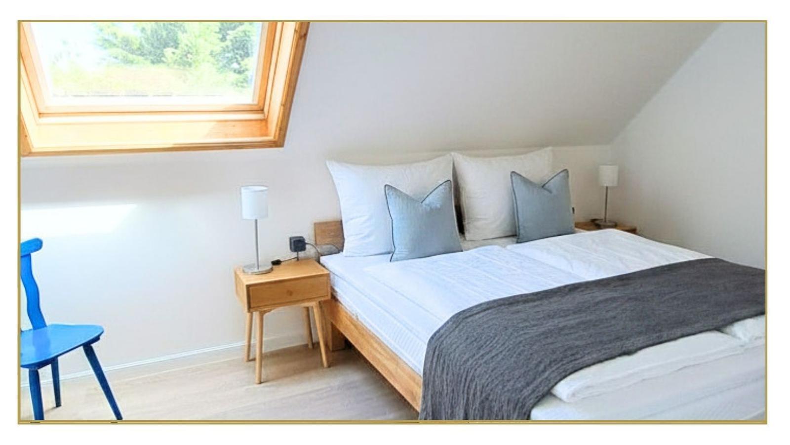 Schlafzimmer mit Dachschrägen, Bett mit Kissen, Nachttisch und blauer Stuhl.