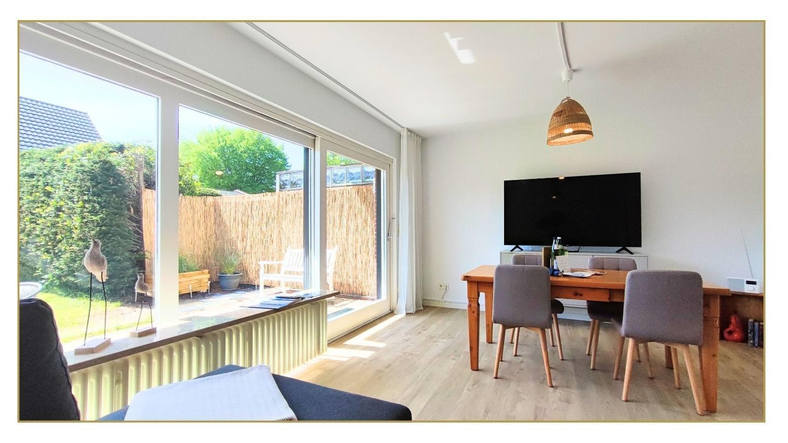Ein Esszimmer mit Holztisch, Stühlen und Fernseher. Große Fenster mit Blick auf einen Garten.