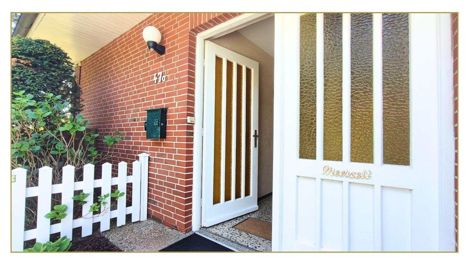 Haus mit rotem Backstein, weißer Tür und weißen Zaun. Hausnummer 47a. Auf der Tür steht Meerzeit.