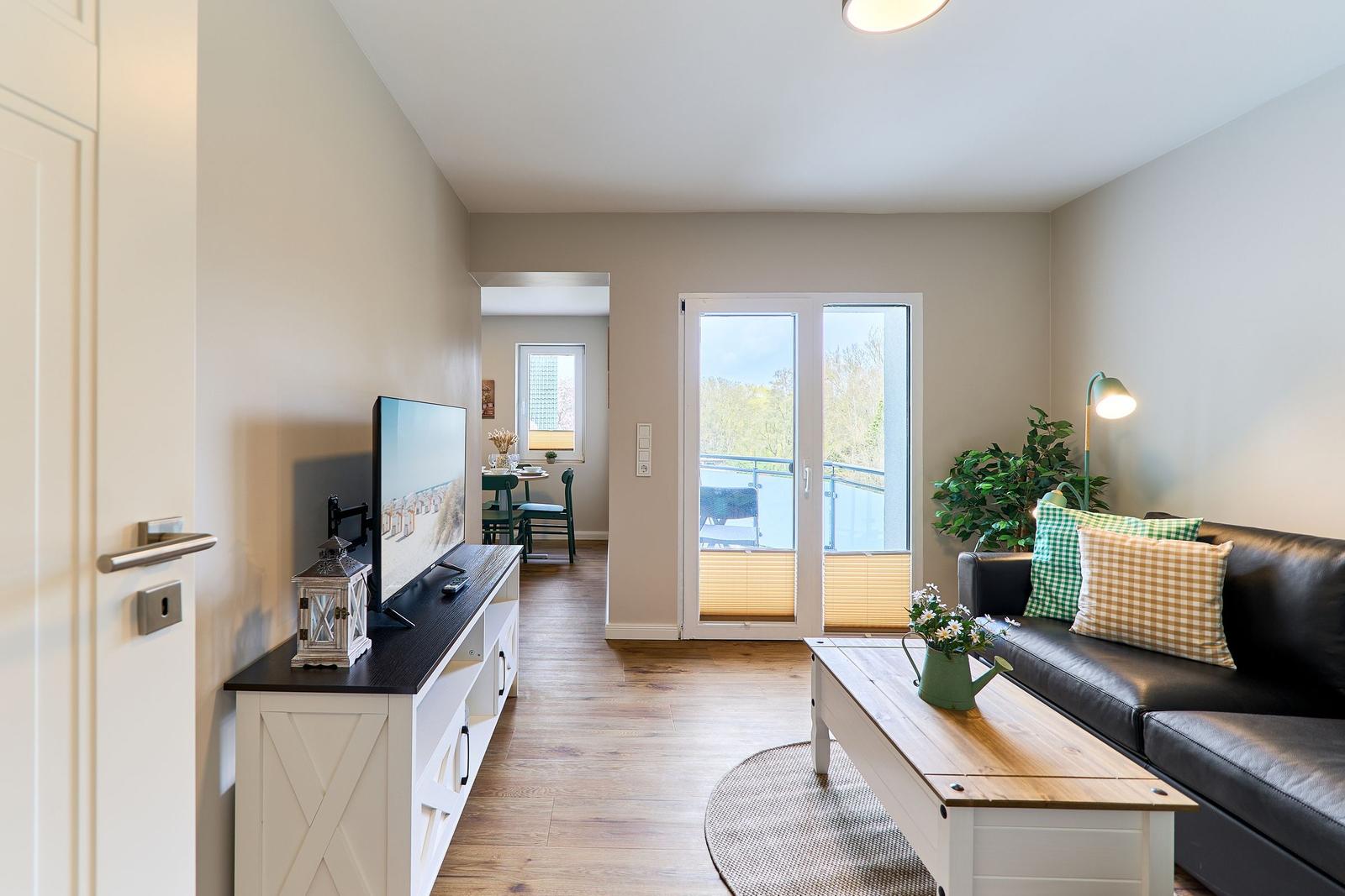 Wohnzimmer mit Sofa, Fernseher und Balkontür. Holzboden und Pflanzen sichtbar.