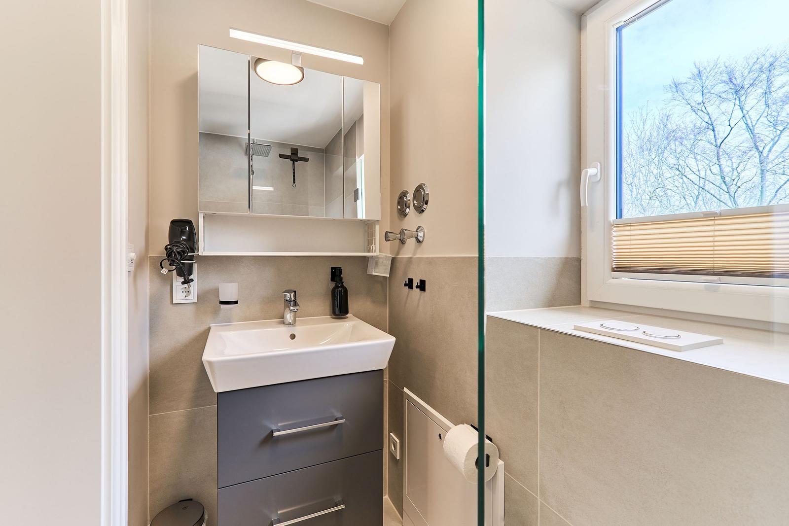 Badezimmer mit Waschbecken, Spiegel und Dusche. Fenster mit Jalousie.