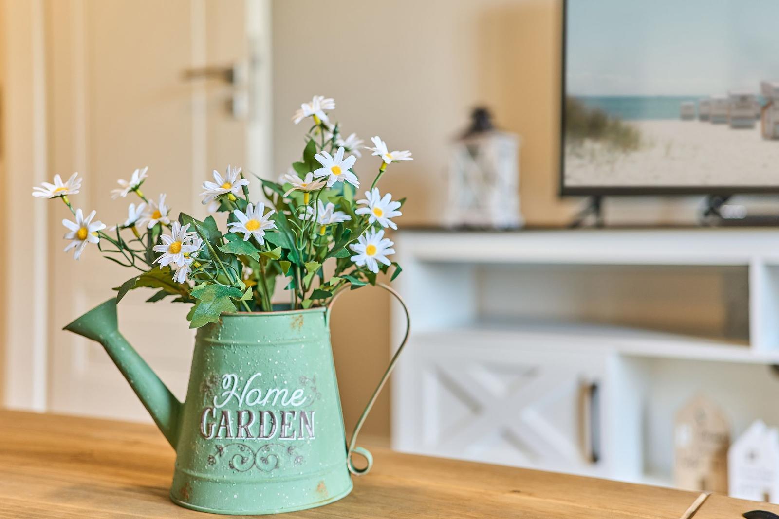 Ein grüner Gießkanister mit Blumen steht auf einem Holztisch. Im Hintergrund ist ein Fernseher zu sehen.