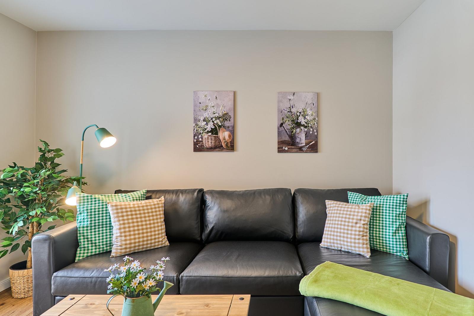 Ein schwarzes Sofa mit Kissen und einem grünen Tuch. Wandbilder und eine Stehlampe sind sichtbar.