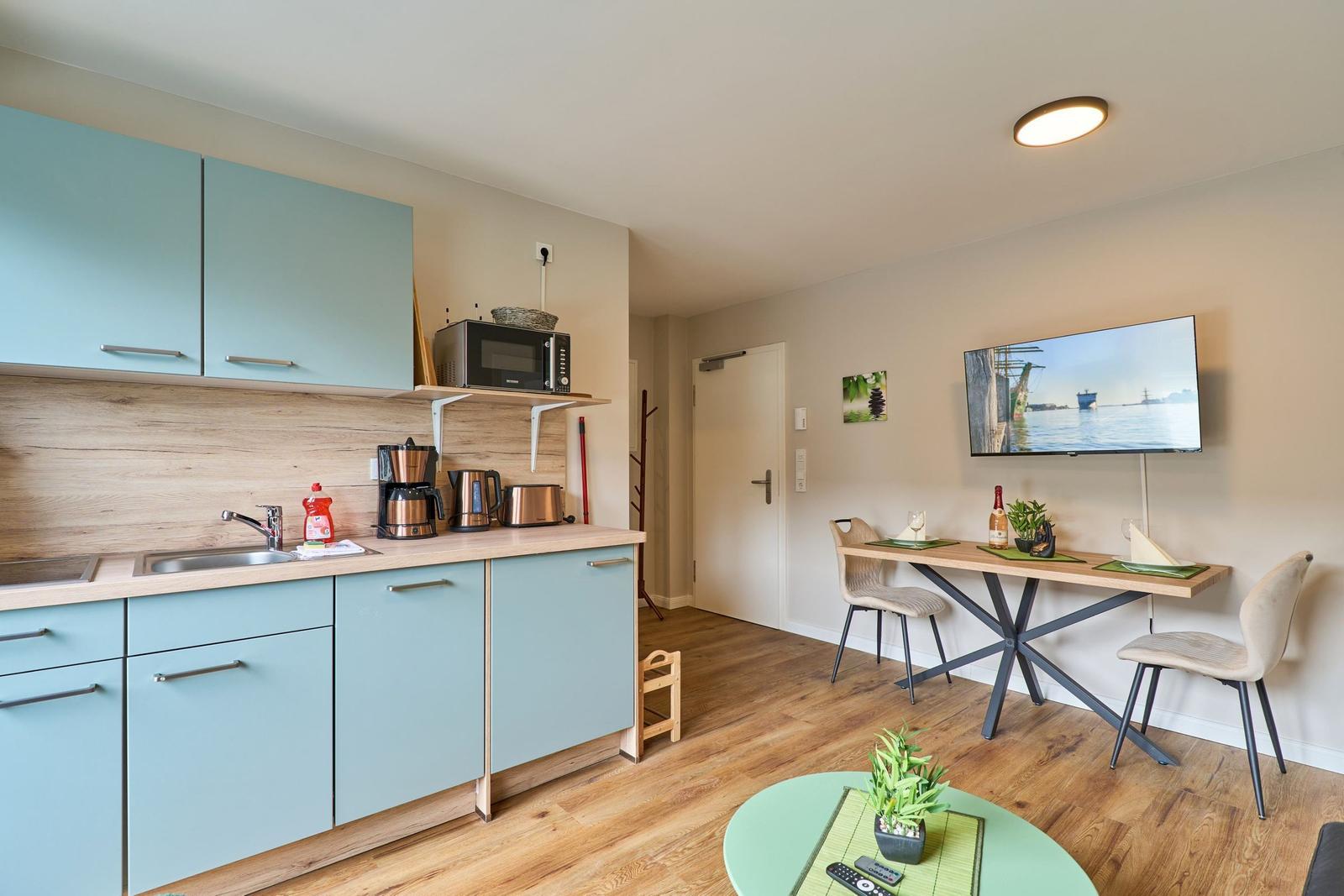 Küche mit Arbeitsplatte, Spüle und Geräten. Essbereich mit Tisch, Stühlen und Fernseher.