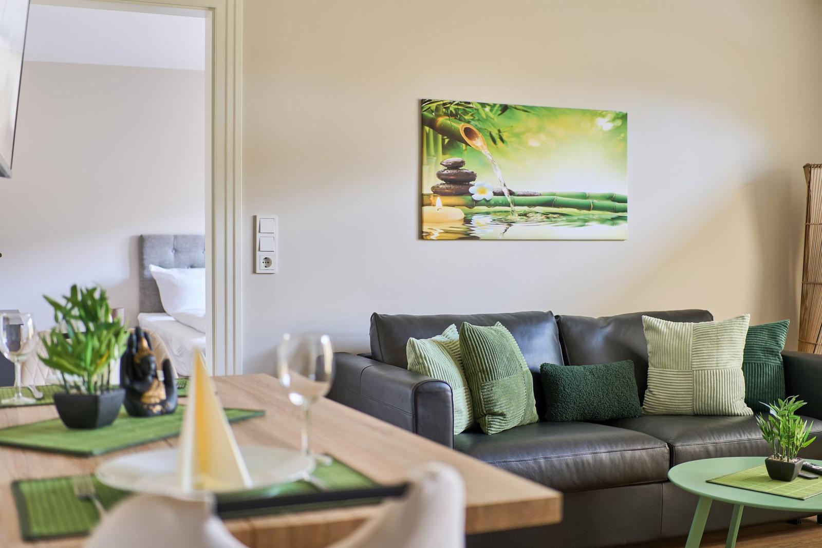 Wohnzimmer mit Sofa, Tisch und Wandbild. Im Hintergrund ist ein Schlafzimmer sichtbar.