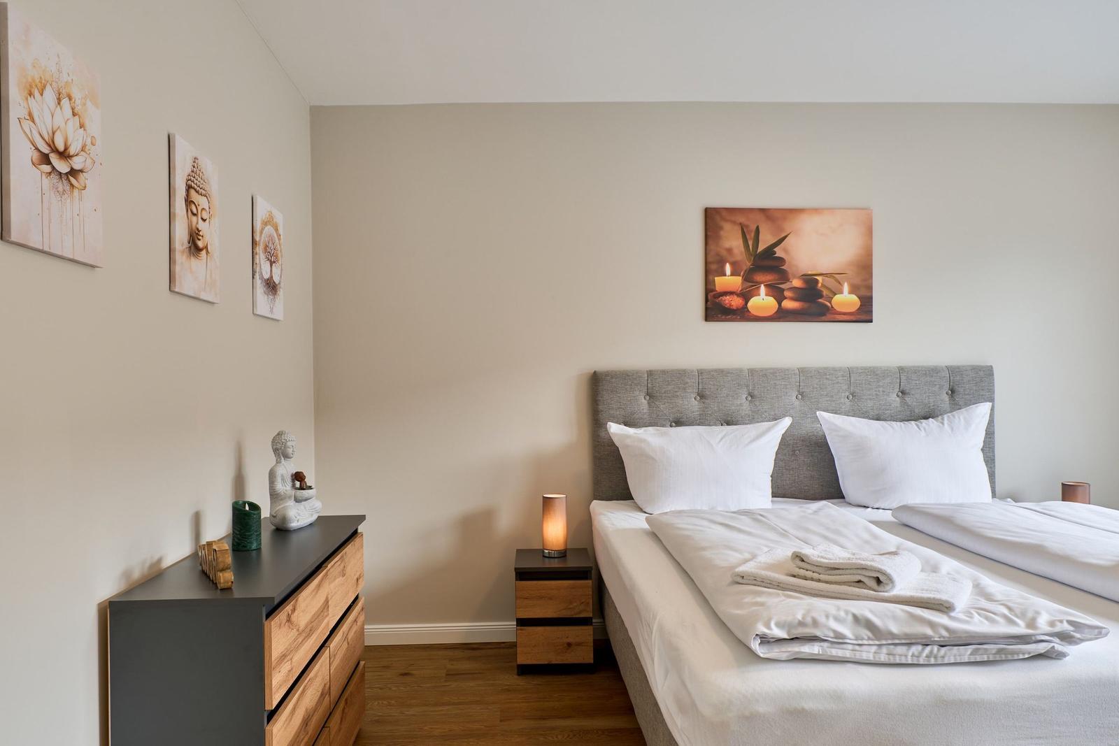 Schlafzimmer mit Bett, Nachttisch und Wandbildern. Holzboden und graue Kopfseite.