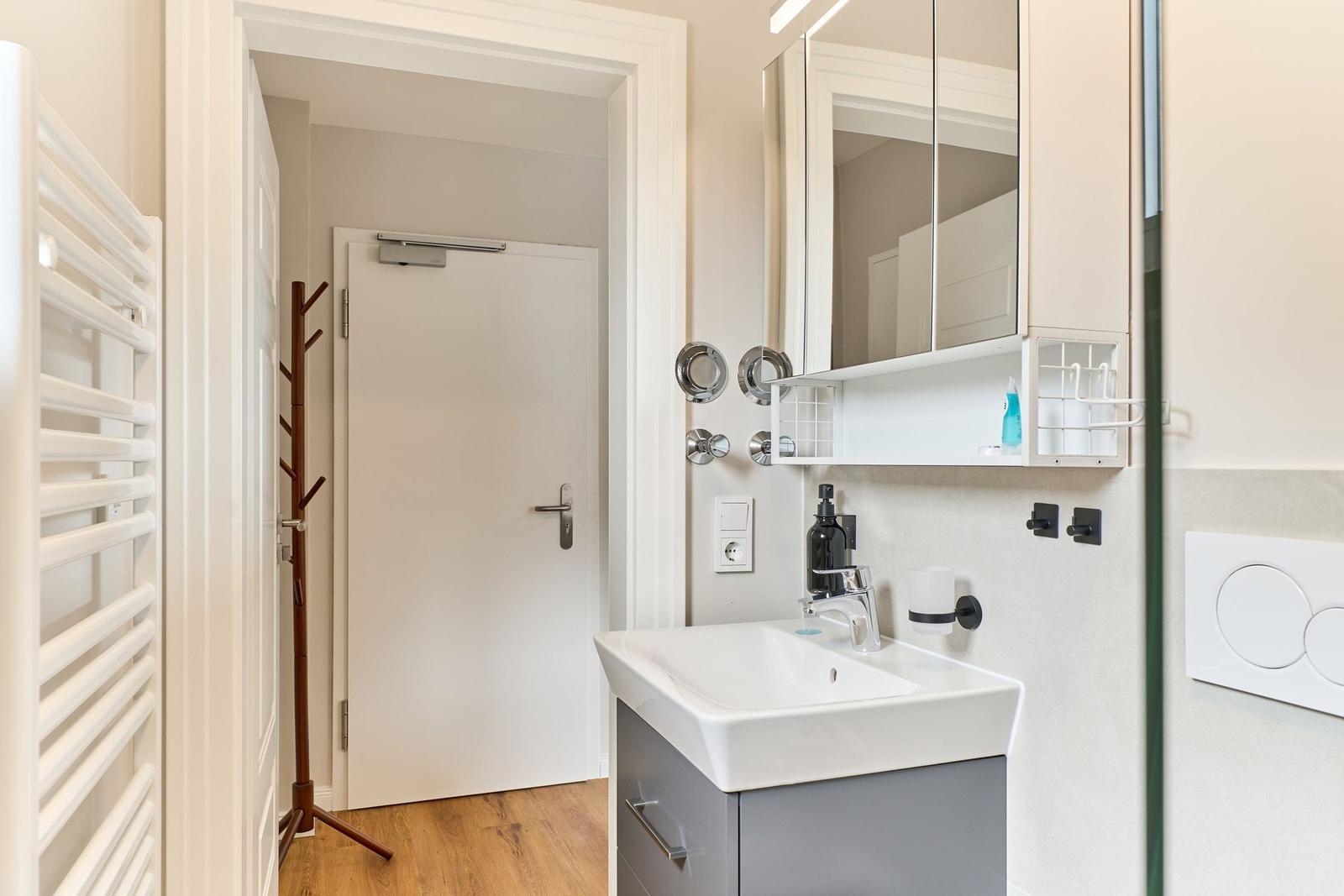 Badezimmer mit Waschbecken, Spiegel und Heizkörper. Tür und Holzboden sind sichtbar.
