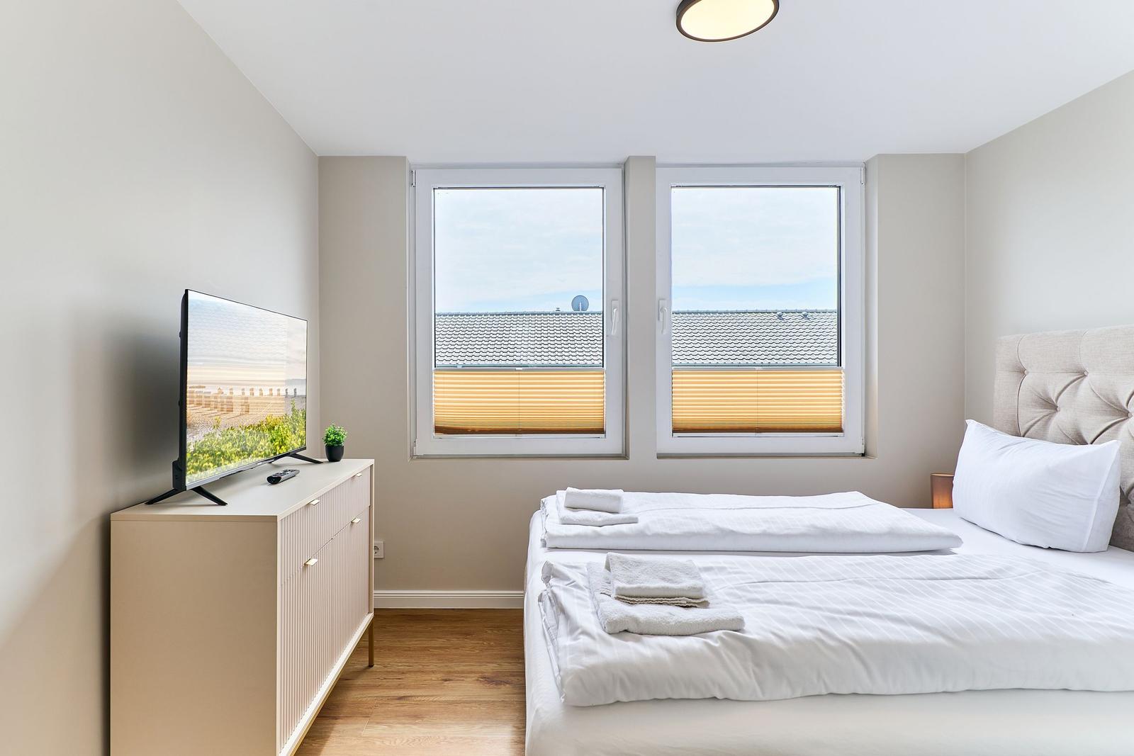 Schlafzimmer mit Bett, Fernseher und Fenster mit Blick auf Dächer.