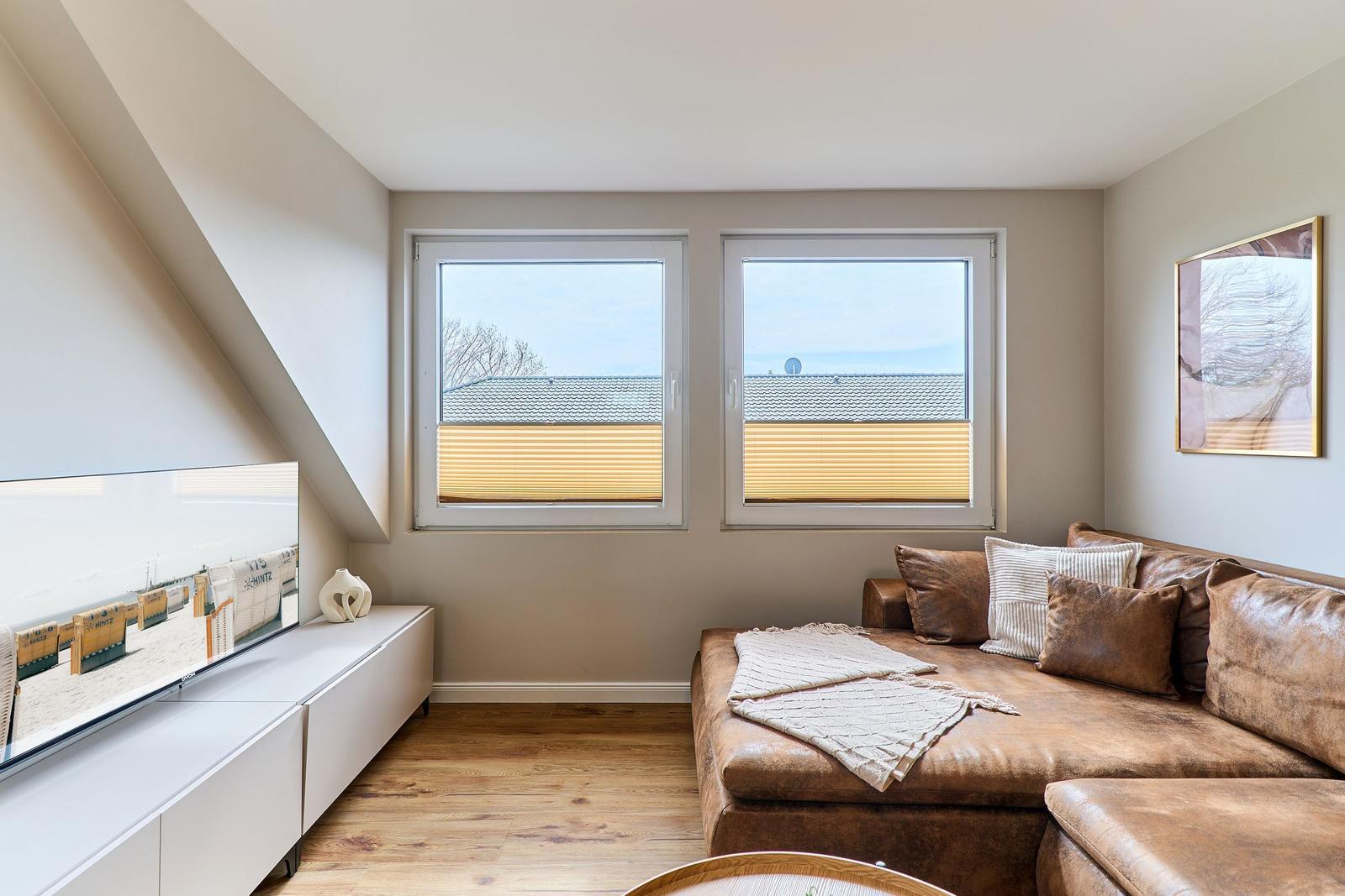 Wohnzimmer mit braunem Sofa, zwei Fenstern und Fernseher auf weißem Sideboard.