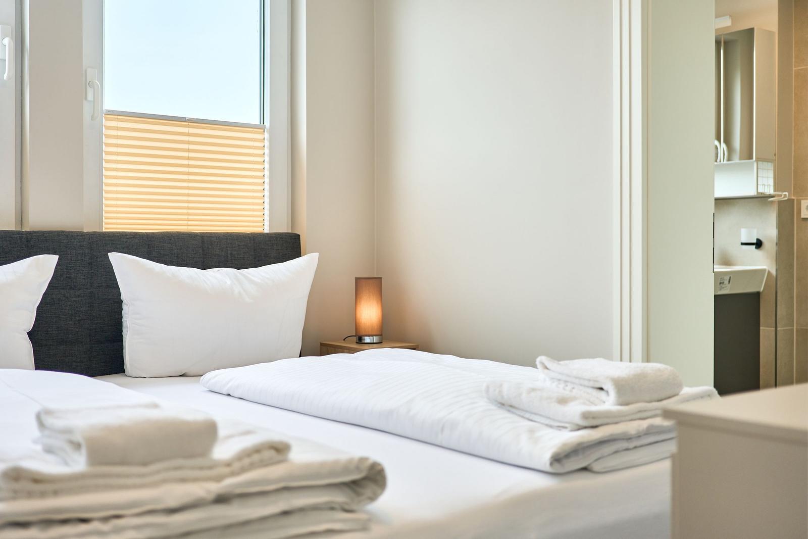 Schlafzimmer mit Bett, Kissen und Falttüchern. Fenster mit Jalousie. Nebenraum mit Waschbecken.