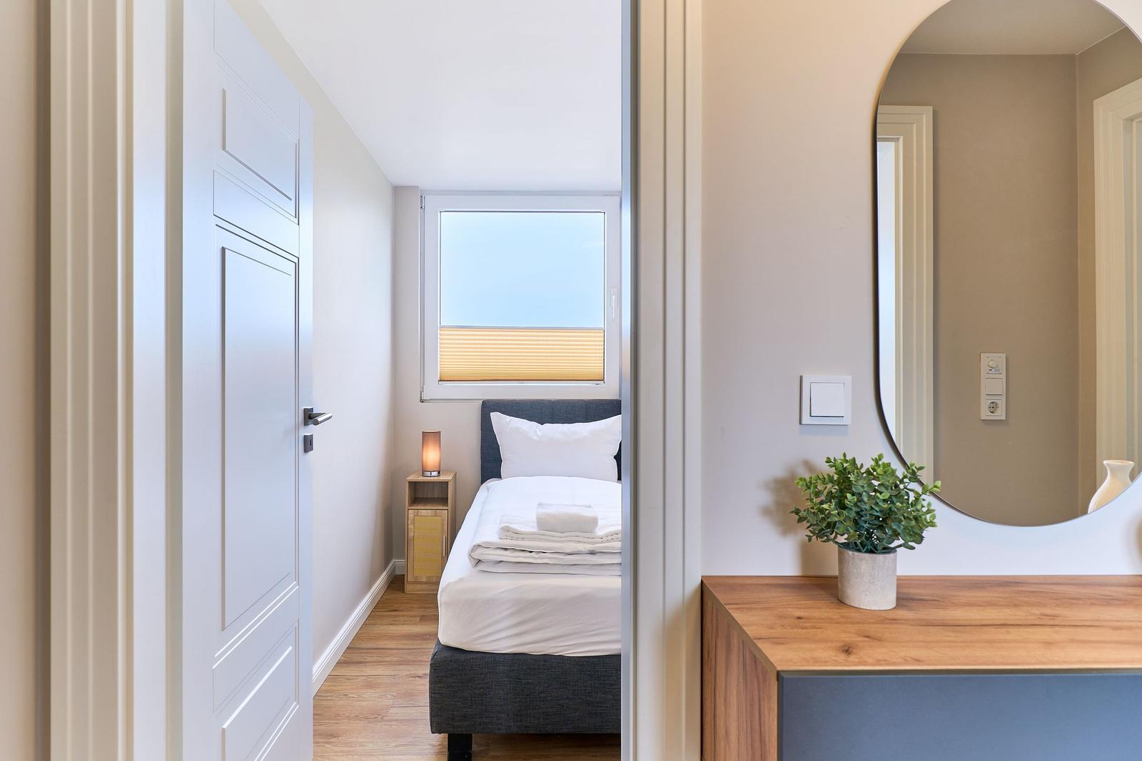 Schlafzimmer mit Bett, Nachttisch und Fenster. Spiegel und Pflanze im Vordergrund.