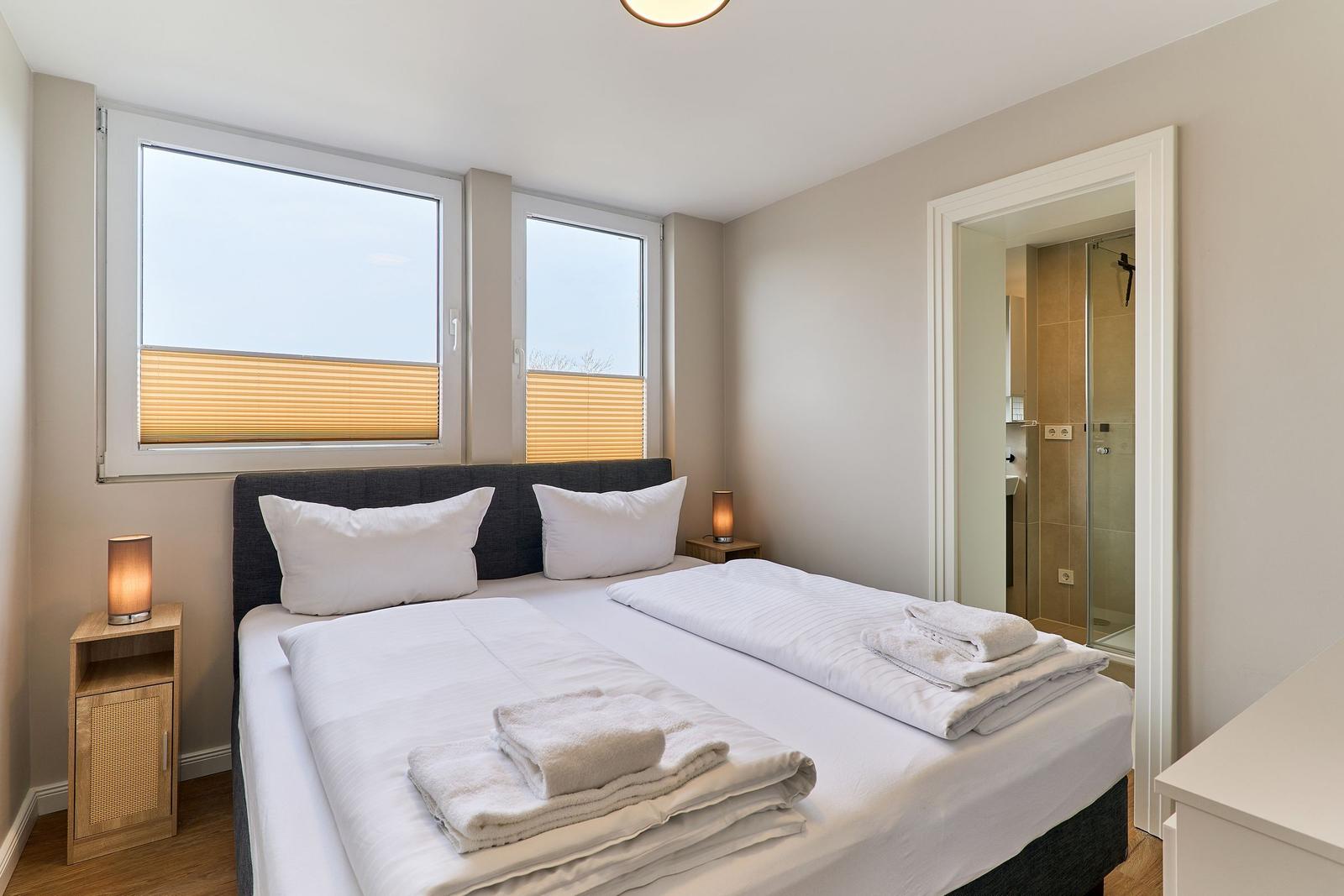 Schlafzimmer mit Doppelbett, zwei Fenstern und Blick in die Dusche.