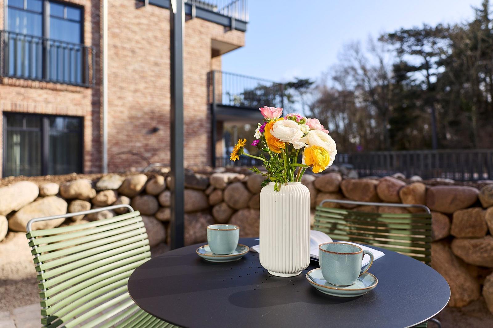 Außenbereich mit Tisch, Stühlen und Blumenarrangement vor Steinmauer und Gebäude