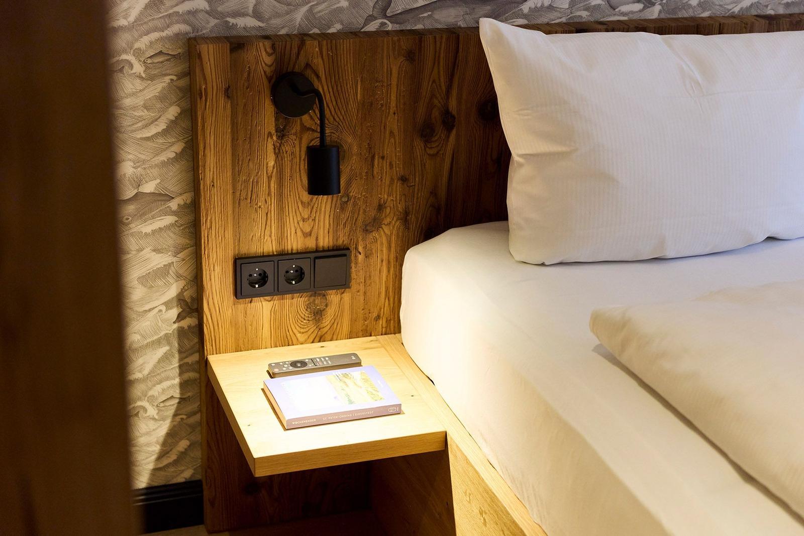 Bettkopfteil aus Holz mit Steckdose und Wandlampe. Nachttisch mit Fernbedienung und Buch.